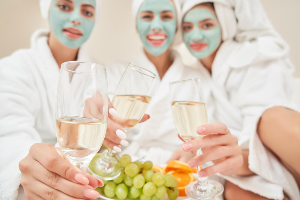 Tre kvinnor i vita morgonrockar som har ansiktsmask och dricker bubbel och ett fruktfat